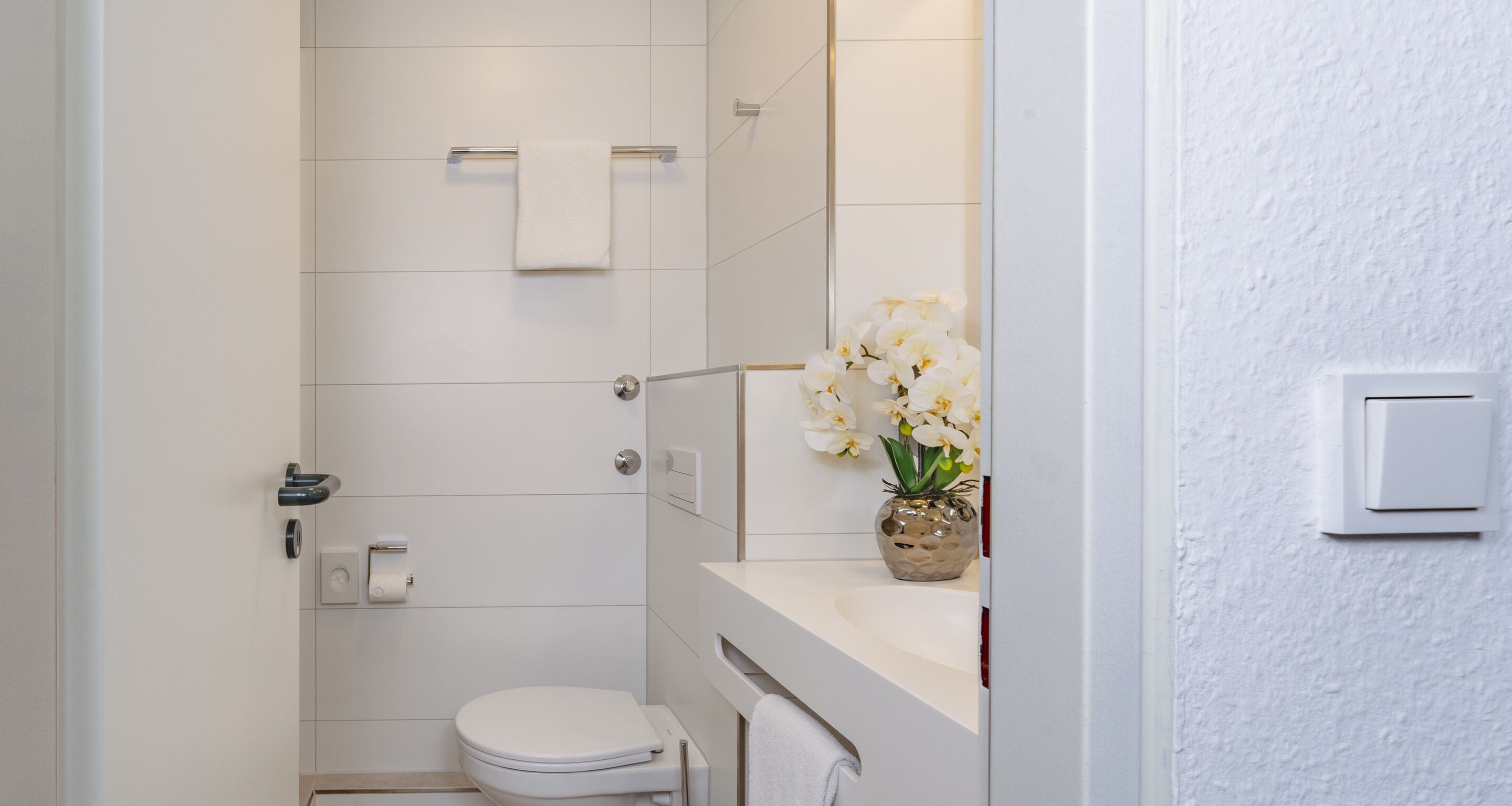 Une salle de bain moderne comprenant des toilettes, un porte-serviettes, un espace douche et un lavabo agrémenté d’un vase de fleurs.