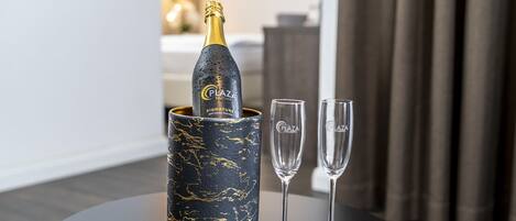 A bottle of Plaza Signature Extra Brut champagne in a black marble-patterned container, accompanied by two champagne flutes on a round table.