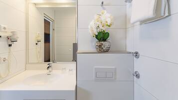 Un baño moderno con un lavabo blanco, un espejo, un jarrón con flores blancas y una toalla colgada en la pared.