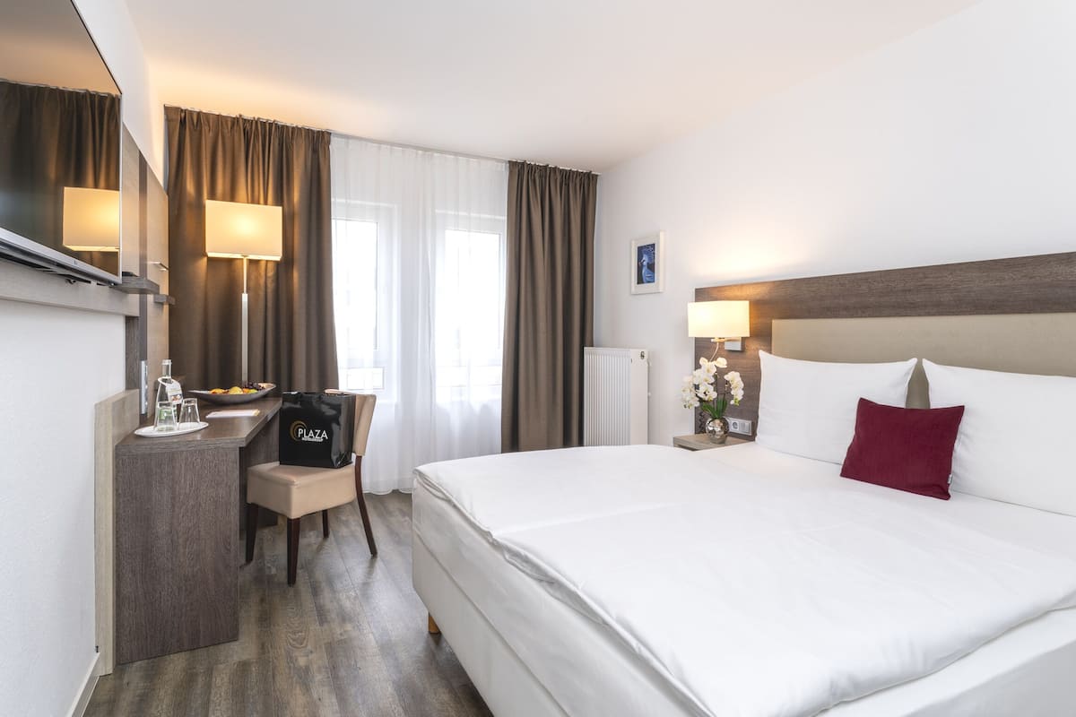 Habitación de hotel con cama, escritorio con silla, lámpara y ventana con cortinas.