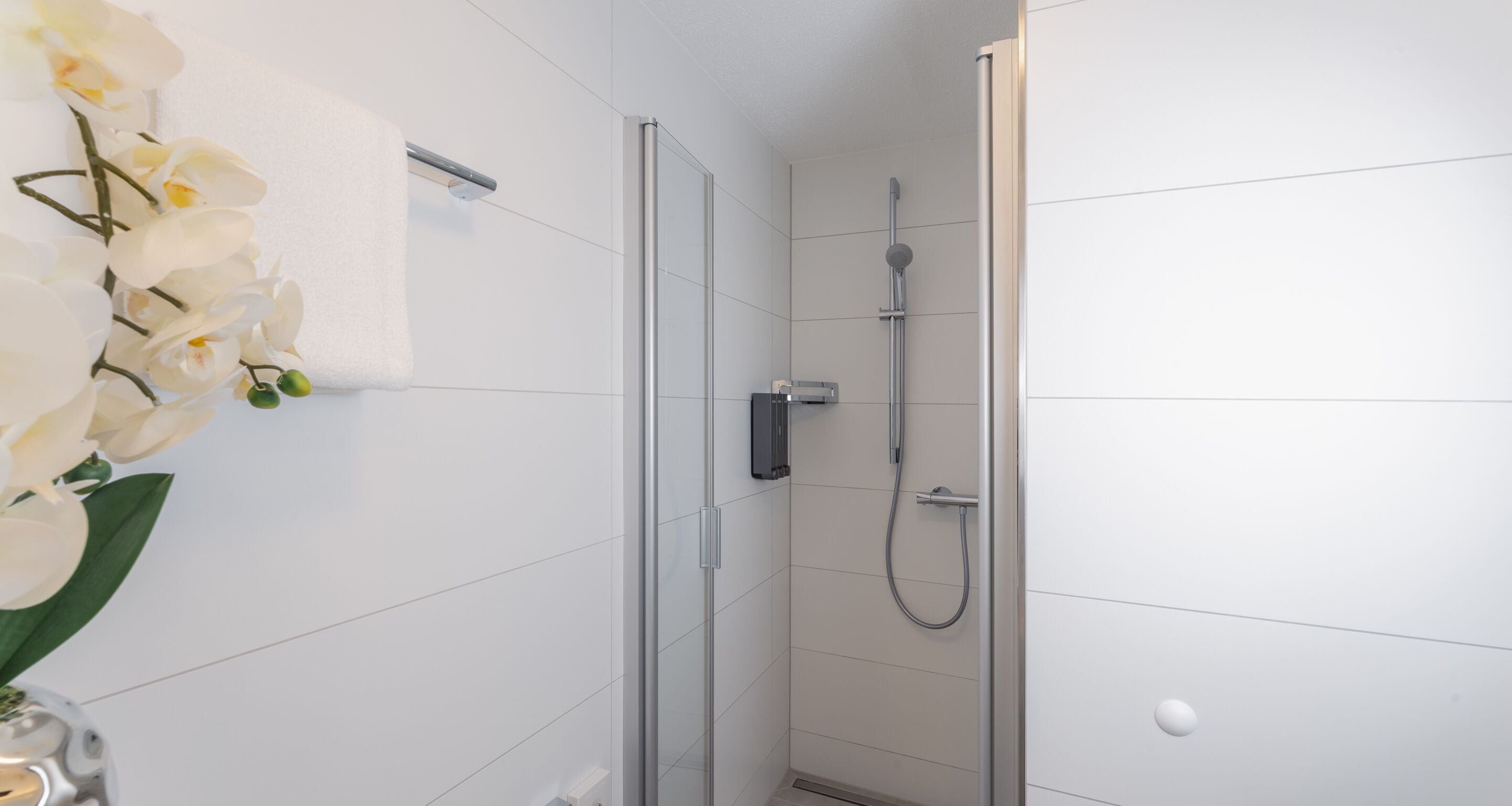 A modern bathroom with white tiled walls, a glass shower enclosure, a handheld showerhead, and a white orchid vase.