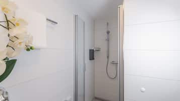 A modern bathroom with white tiled walls, a glass shower enclosure, a handheld showerhead, and a white orchid vase.