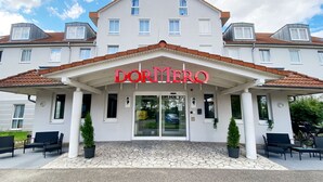 Een hotelingang met een bord waarop "DORMERO" staat en een overdekt entreegebied met stoelen en plantenpotten.