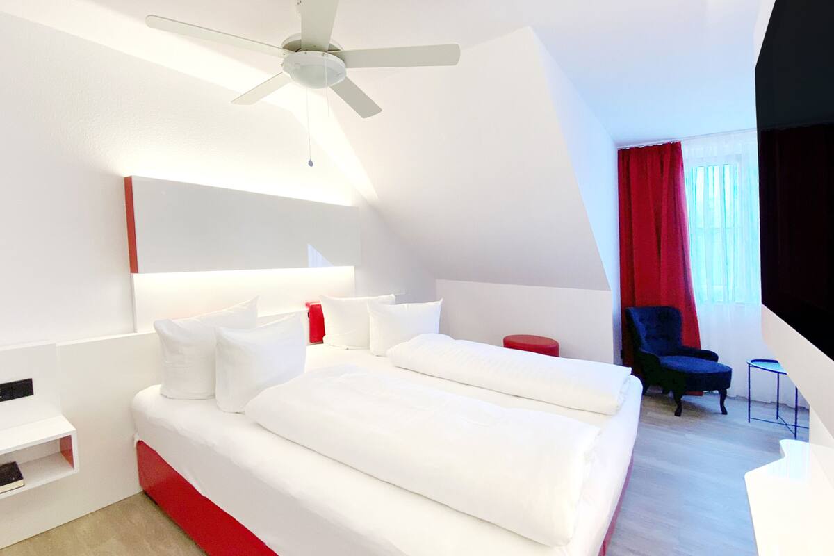 Ein modernes Hotelzimmer mit einem großen Bett, einem Deckenventilator, einer roten Akzentwand, einem roten Sessel und einem kleinen Tisch.