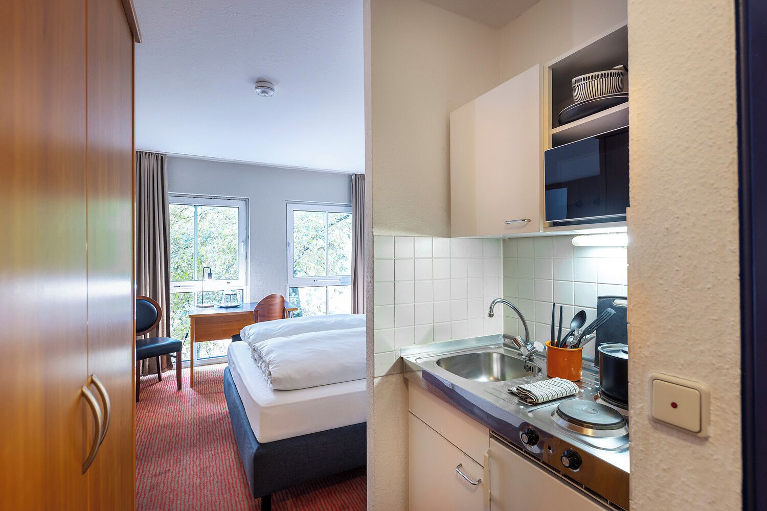Una habitación de hotel con una cama, un escritorio, una silla, una ventana y una kitchenette.