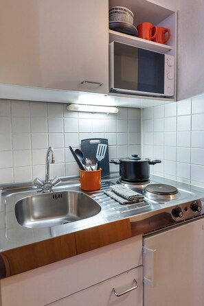 A kitchen with a stainless steel sink, a stove, a microwave, and a shelf with kitchenware. - ACHAT Hotel Darmstadt Griesheim (Griesheim)