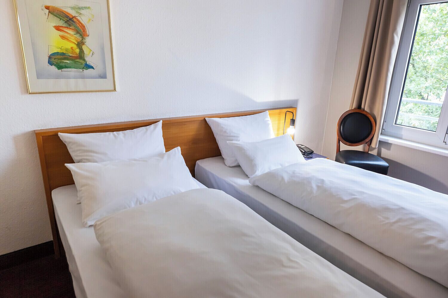 Une chambre d’hôtel avec deux lits simples, une tête de lit en bois, une table de chevet avec une lampe, une chaise et une fenêtre avec des rideaux.