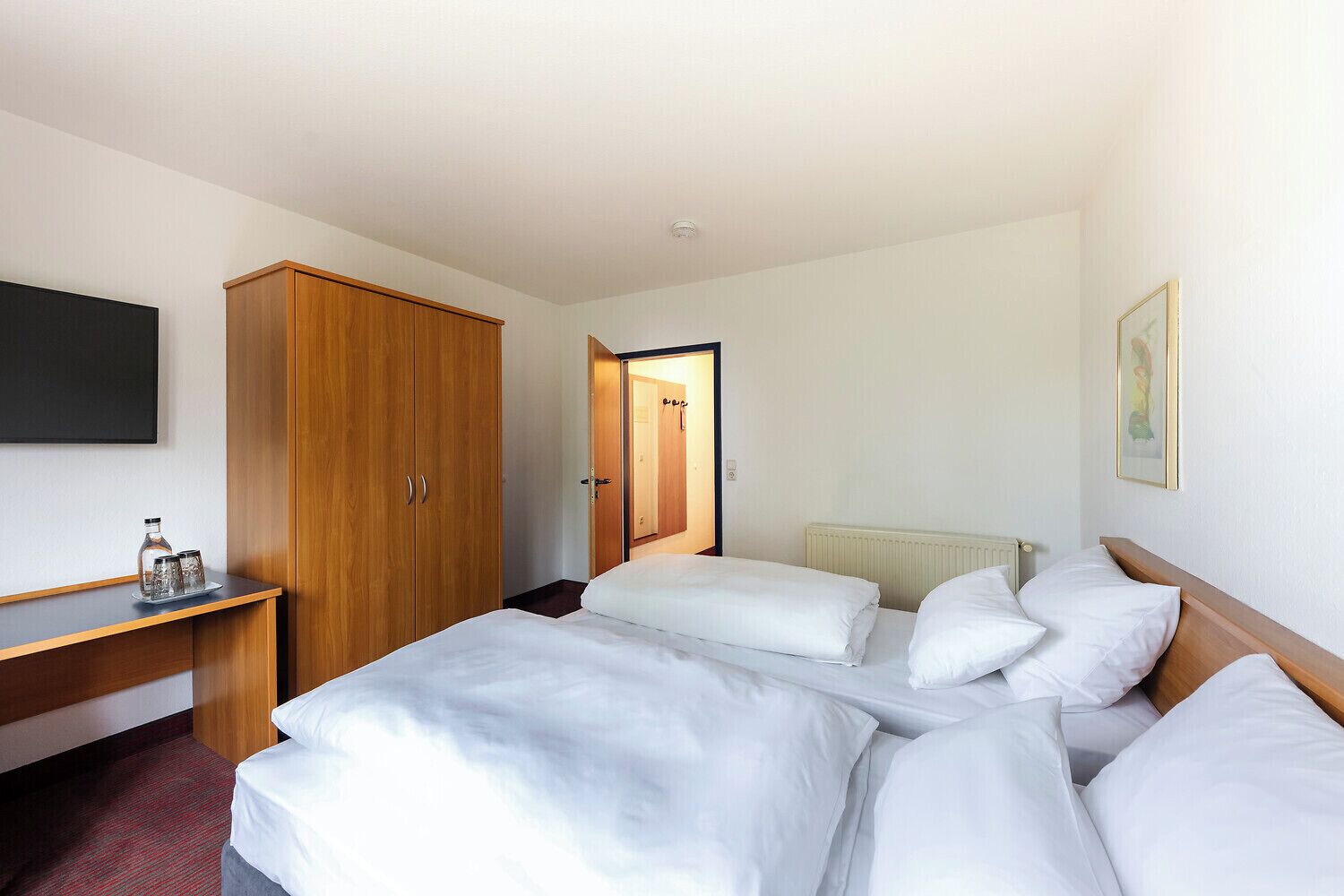 Habitación de hotel con cama, armario, mesita de noche con vasos y un cuadro en la pared.