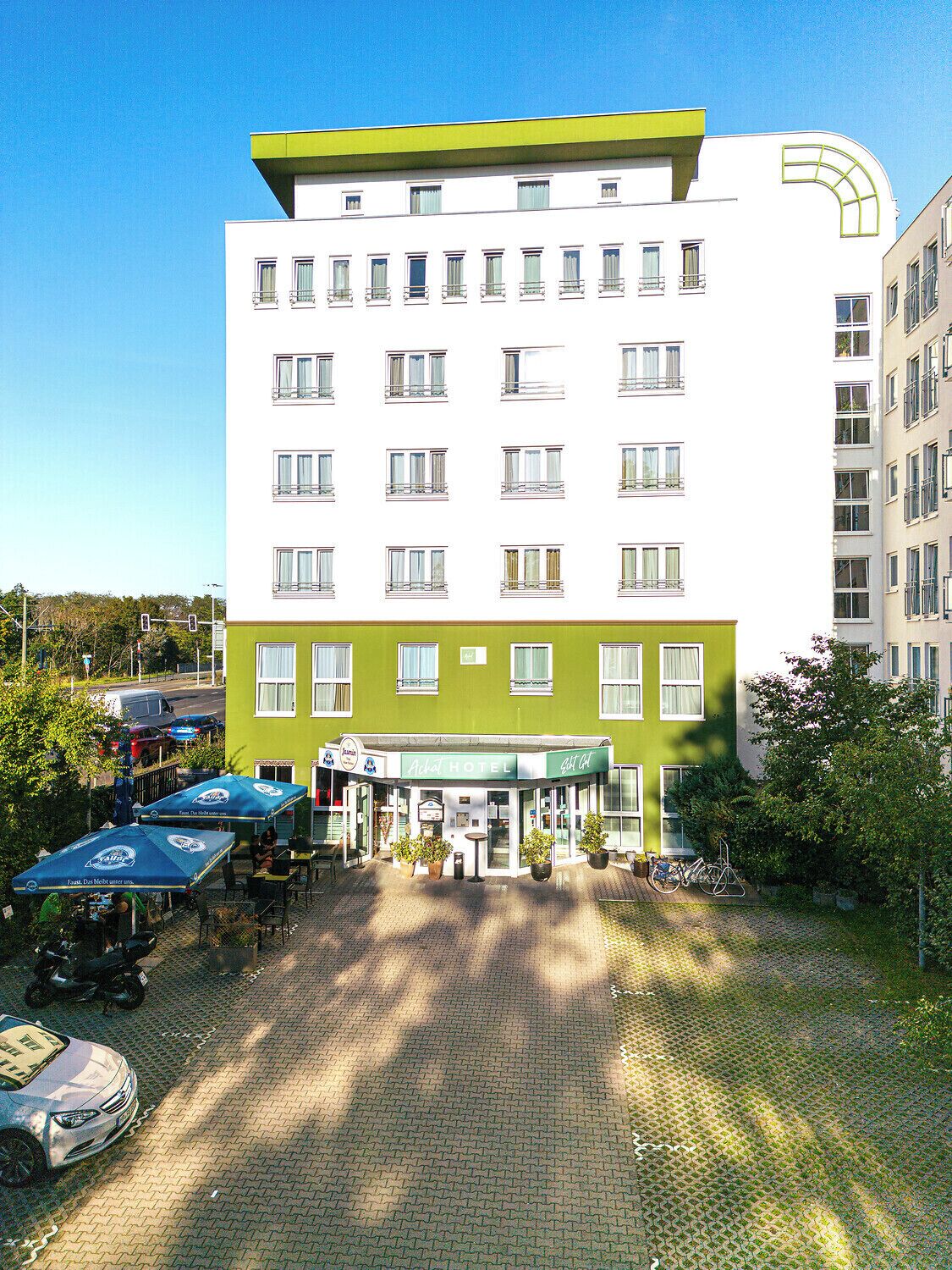 A hotel with a green and white facade, multiple windows, and a covered outdoor seating area with umbrellas.