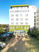 Um hotel com fachada verde e branca, várias janelas e uma área de estar coberta ao ar livre com guarda-sóis.