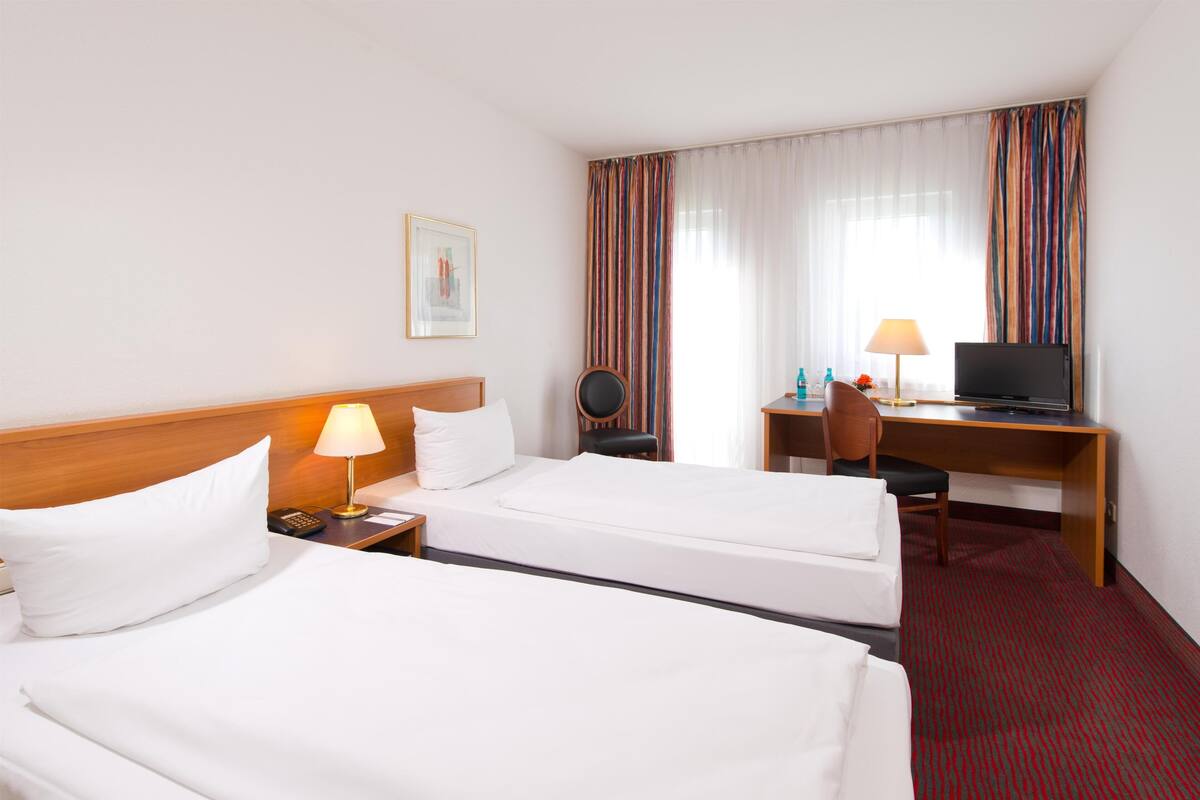 A hotel room with two beds, a desk, a chair, a TV, and a lamp.