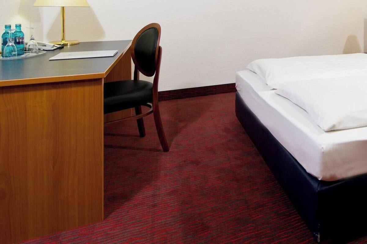 A hotel room with a bed, a desk, a chair, and a nightstand with a lamp and a flower.