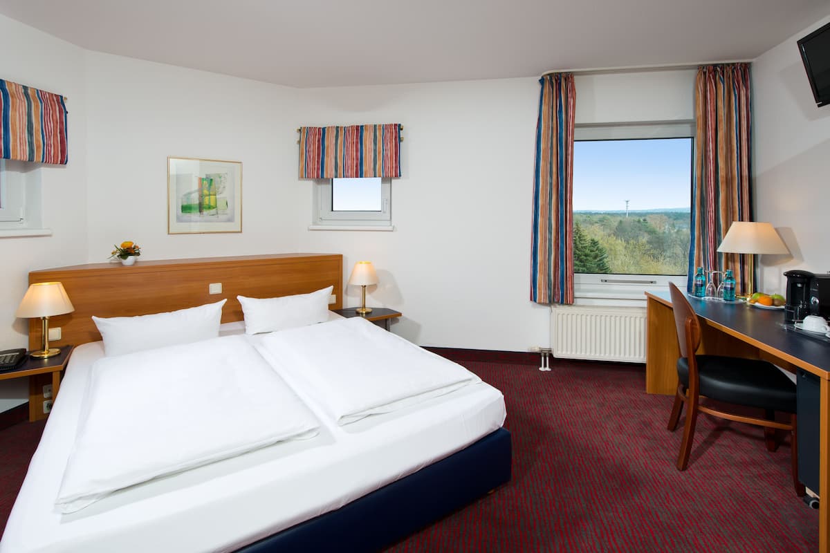 A hotel room with a large bed, two bedside tables with lamps, a desk with a chair, a window with curtains, and a framed picture on the wall.