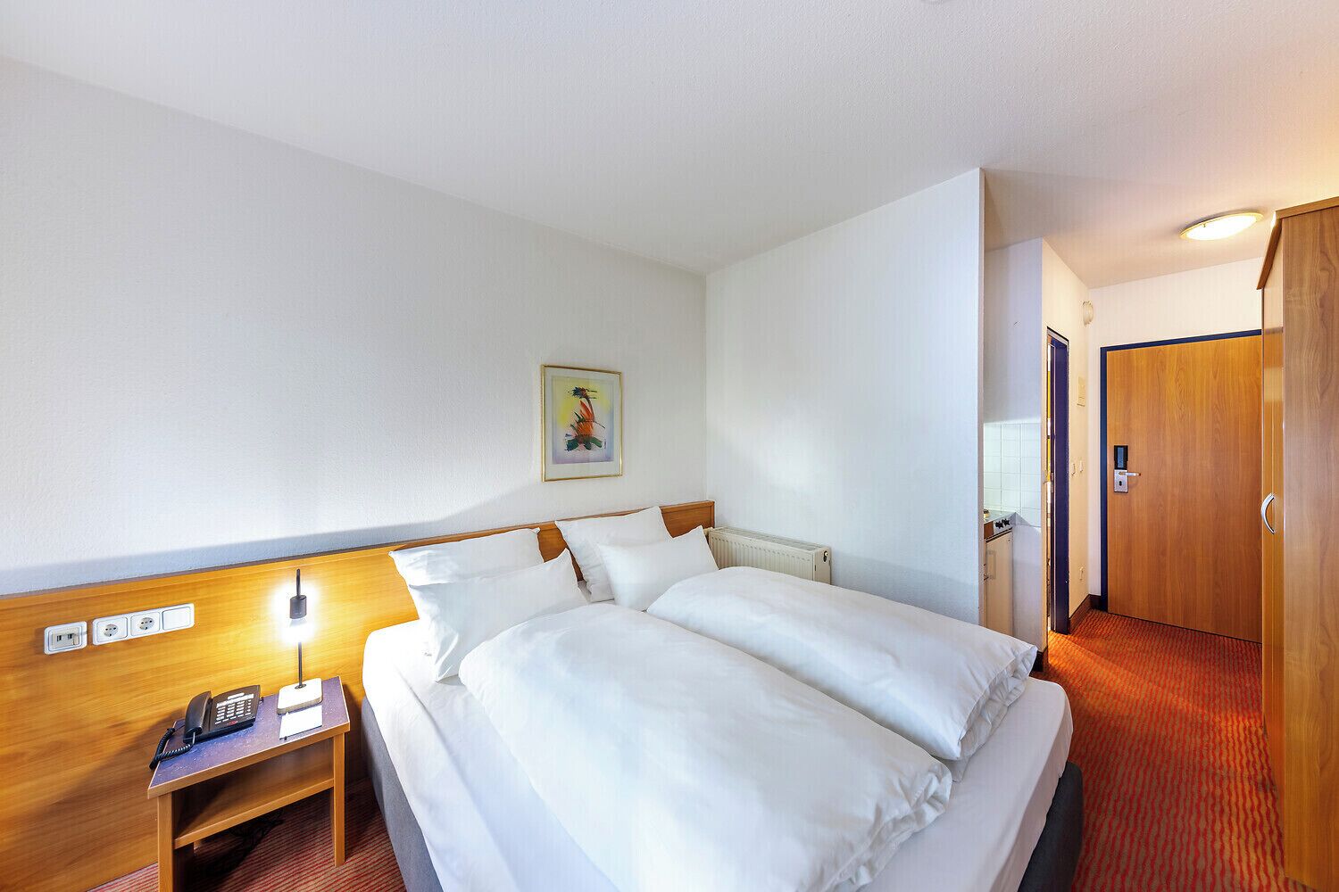 Ein Hotelzimmer mit Doppelbett, Nachttisch mit Telefon, Heizkörper, Holzschrank und einer Tür zu einem weiteren Raum.