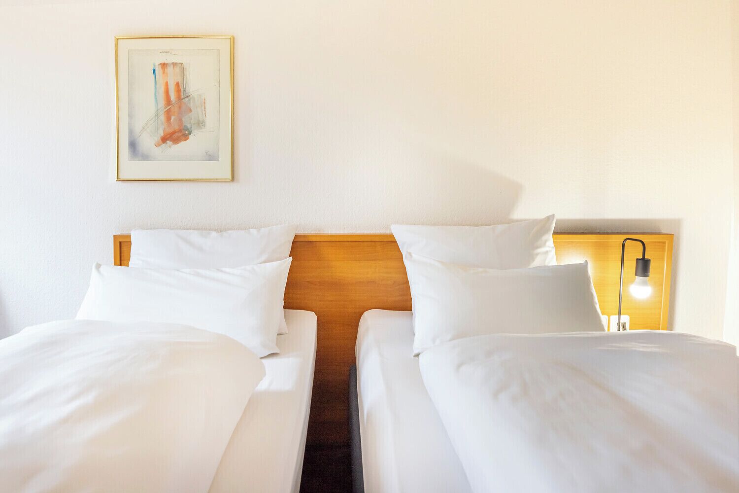 A hotel room with two single beds, a wooden headboard, a bedside lamp, and a framed artwork on the wall.
