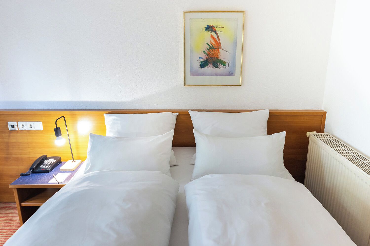 A hotel room with a double bed, a bedside table with a lamp, a phone, and a painting on the wall.