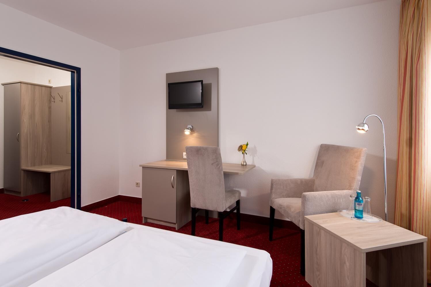 A hotel room with a bed, a desk, two chairs, a TV, a lamp, and a small table with a vase of flowers.
