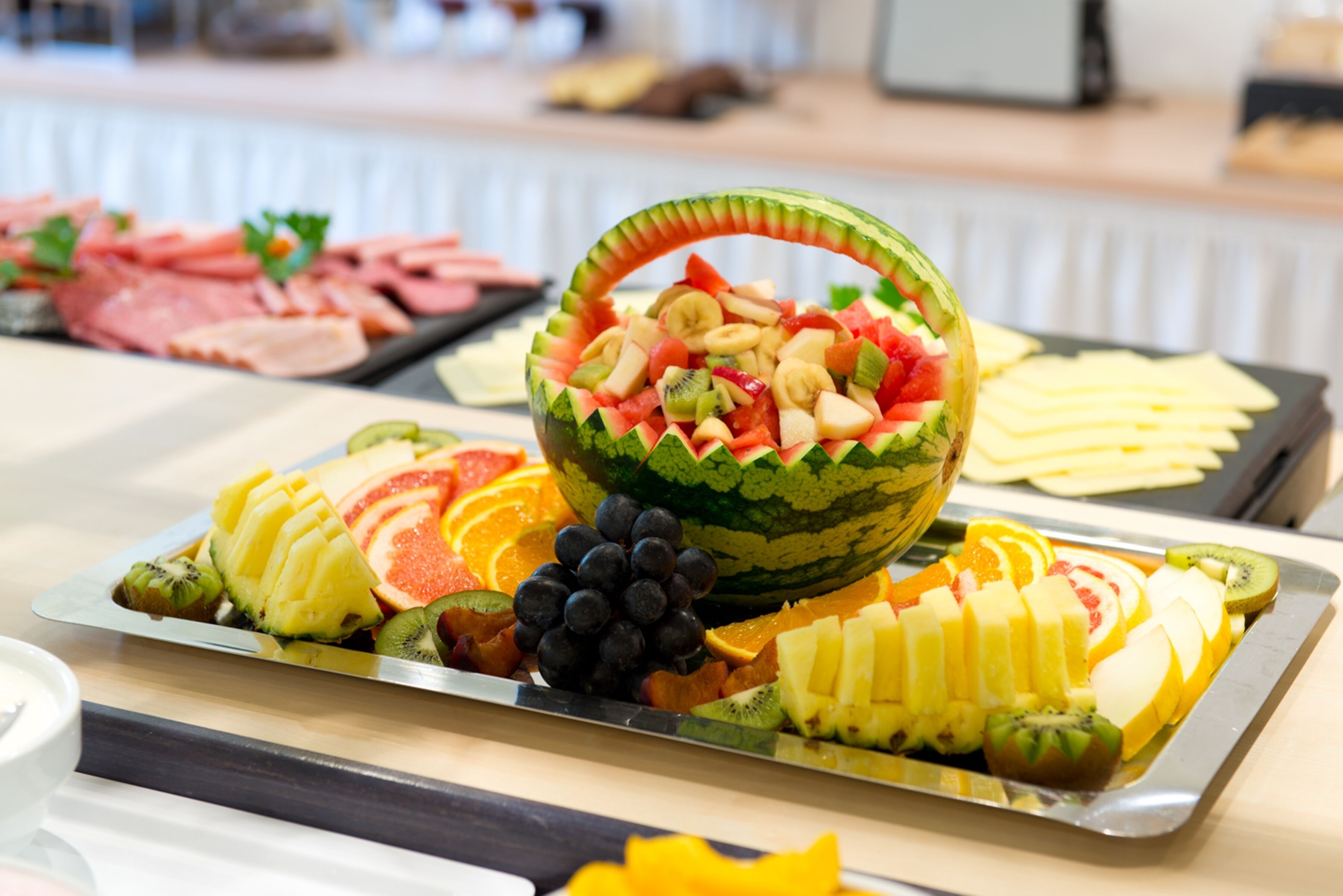 Un plateau de fruits avec un melon creusé servant de récipient, contenant des raisins, des kiwis et des morceaux de melon, le tout entouré de fruits coupés en tranches et de fromage, le tout disposé sur un plateau.