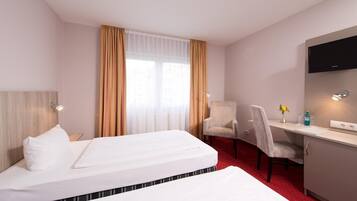 A hotel room with a bed, a desk, two chairs, a TV, and a window with curtains.