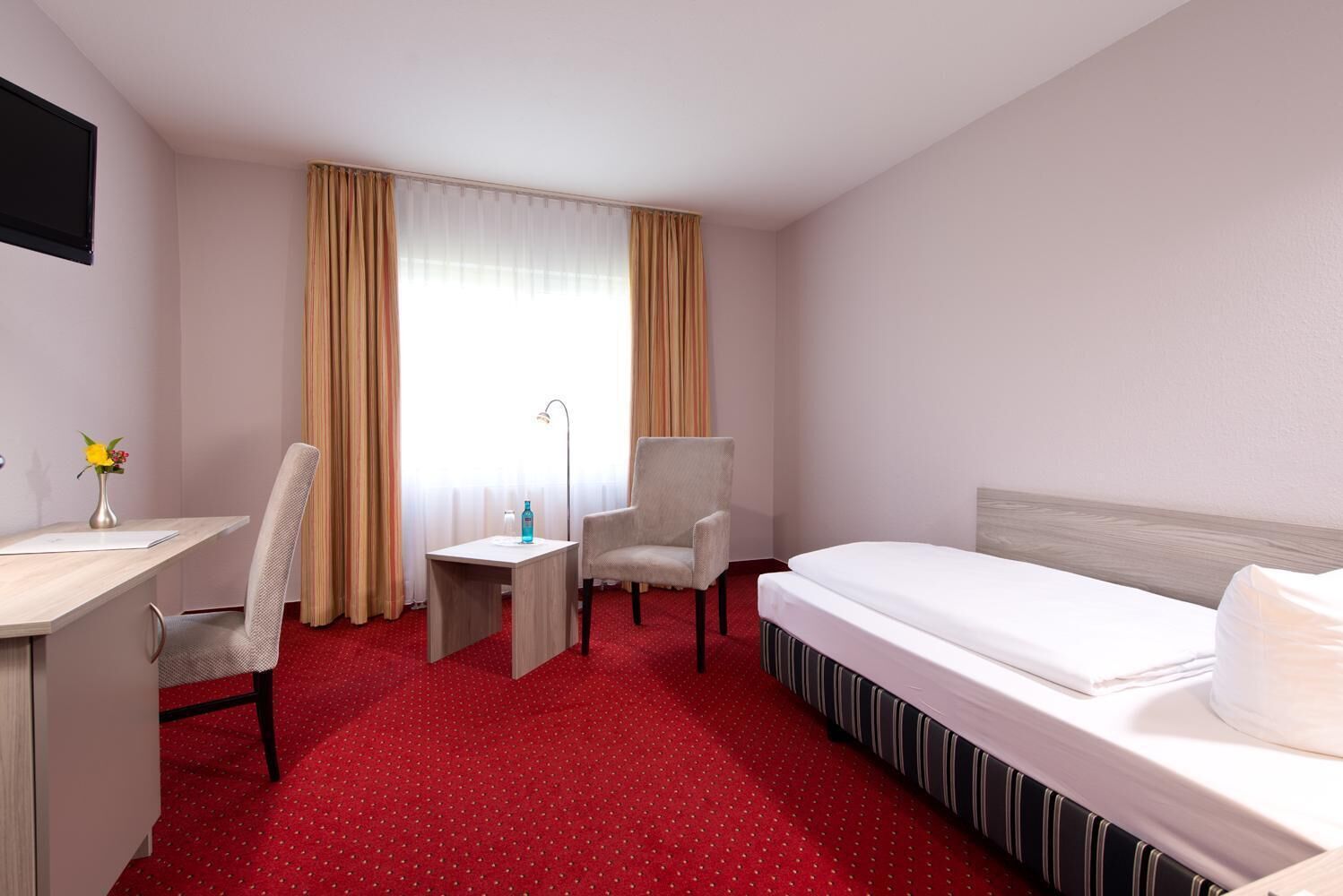 Ein Hotelzimmer mit einem Bett, zwei Stühlen, einem Schreibtisch, einer Lampe und einem fenster mit Vorhängen.