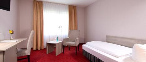 Een hotelkamer met een bed, twee stoelen, een bureau, een lamp en een raam met gordijnen.