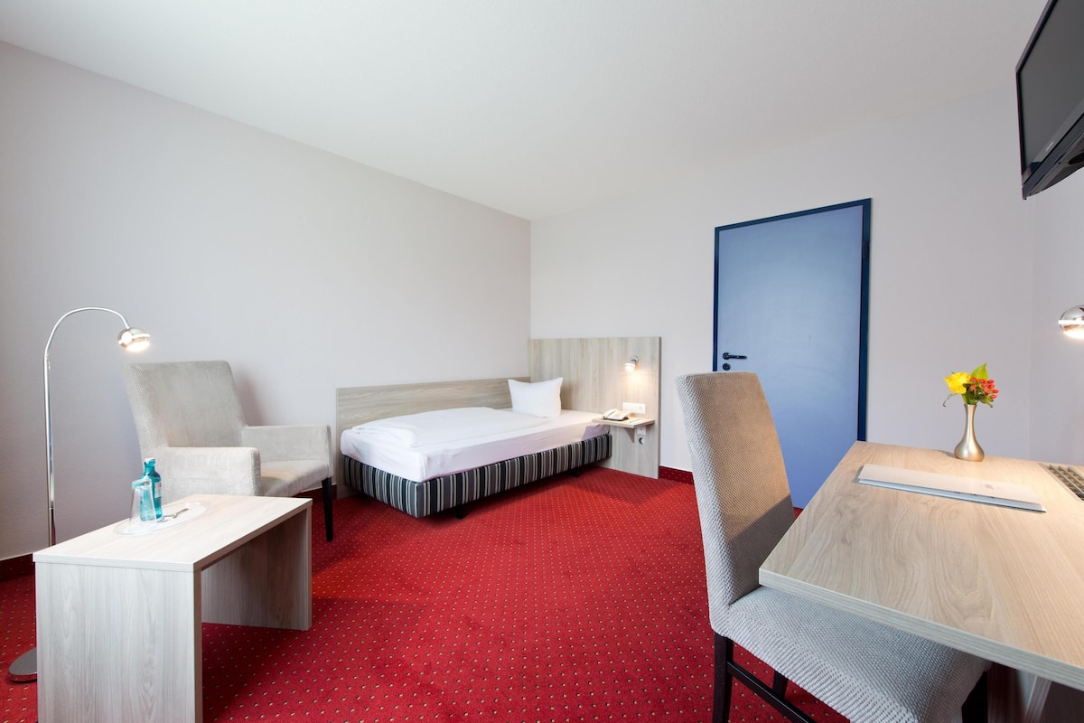 Una habitación de hotel con una cama, dos sillas, un escritorio, una lámpara y un florero con flores.
