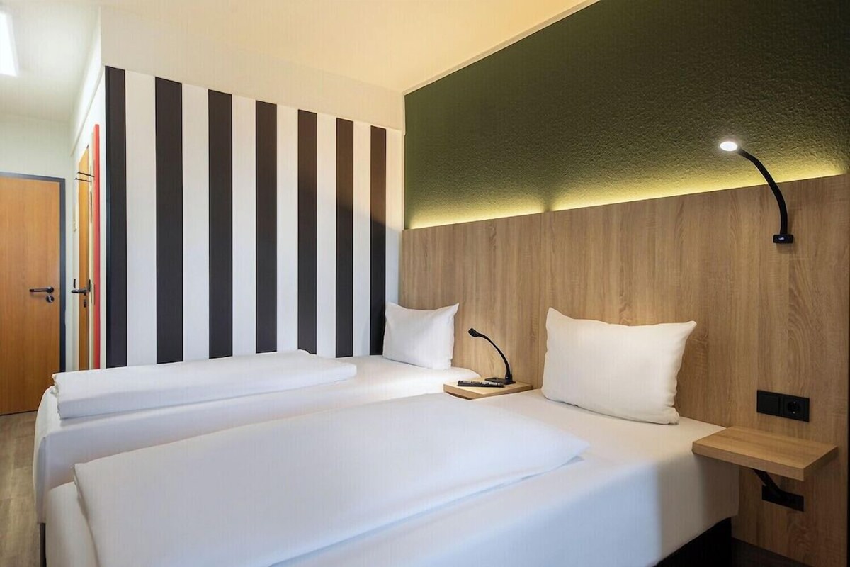 A hotel room with two beds, a wooden headboard, a bedside table with a lamp, and a striped wall.