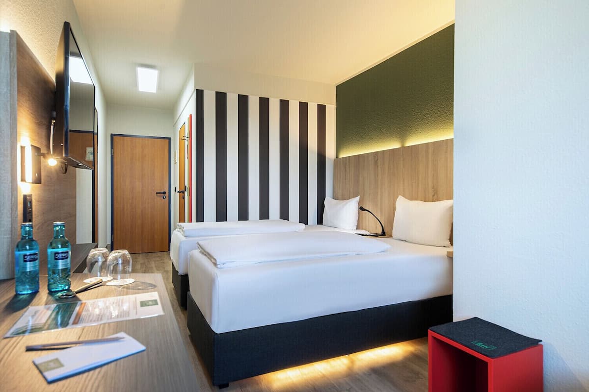 Quarto de hotel com duas camas, uma televisão, uma escrivaninha com cadeira e uma mesa de apoio vermelha.