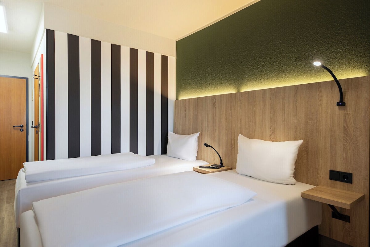 Quarto de hotel com duas camas, parede listrada, cabeceira de madeira e mesa de cabeceira com abajur.