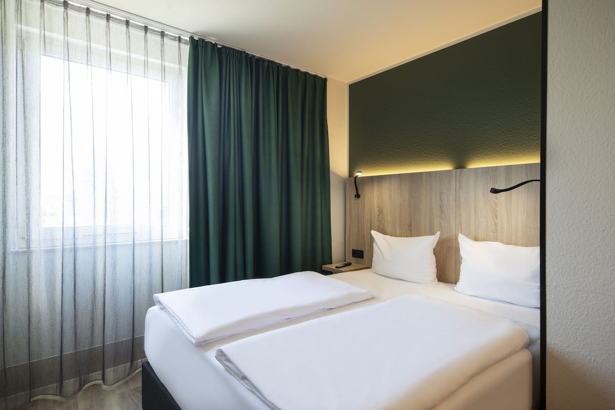 Quarto de hotel com duas camas, cabeceira de madeira, mesa de cabeceira e janela com cortinas finas.