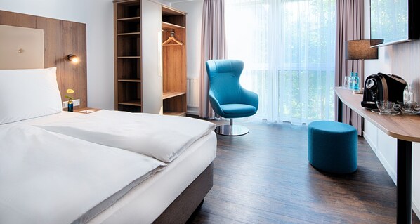 Habitación de hotel con cama, una silla azul, un pequeño otoman, un escritorio con lámpara y un armario.
