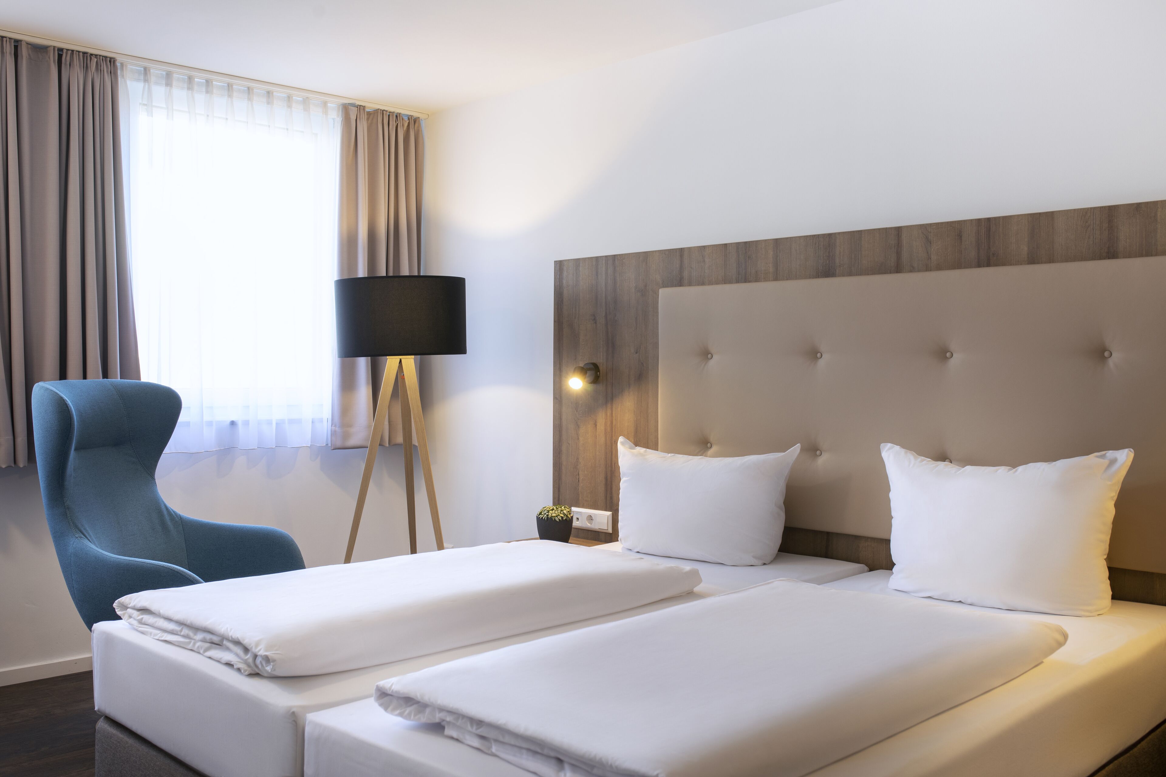 Une chambre d’hôtel avec deux lits, un fauteuil bleu, une lampe sur pied et une fenêtre avec des rideaux.