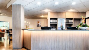 Un mostrador de recepción moderno con acabado en madera clara, una planta grande con flores y un piso de baldosas limpias.