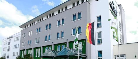 Un hotel de varios pisos con diseño moderno, que presenta fachada verde y blanca, múltiples ventanas y una destacada exhibición de banderas.
