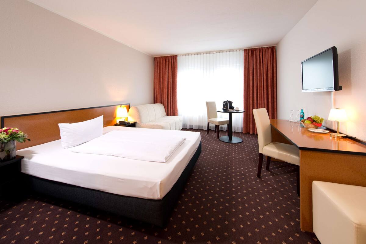 A hotel room with a bed, a desk, a chair, a TV, and a window with curtains.