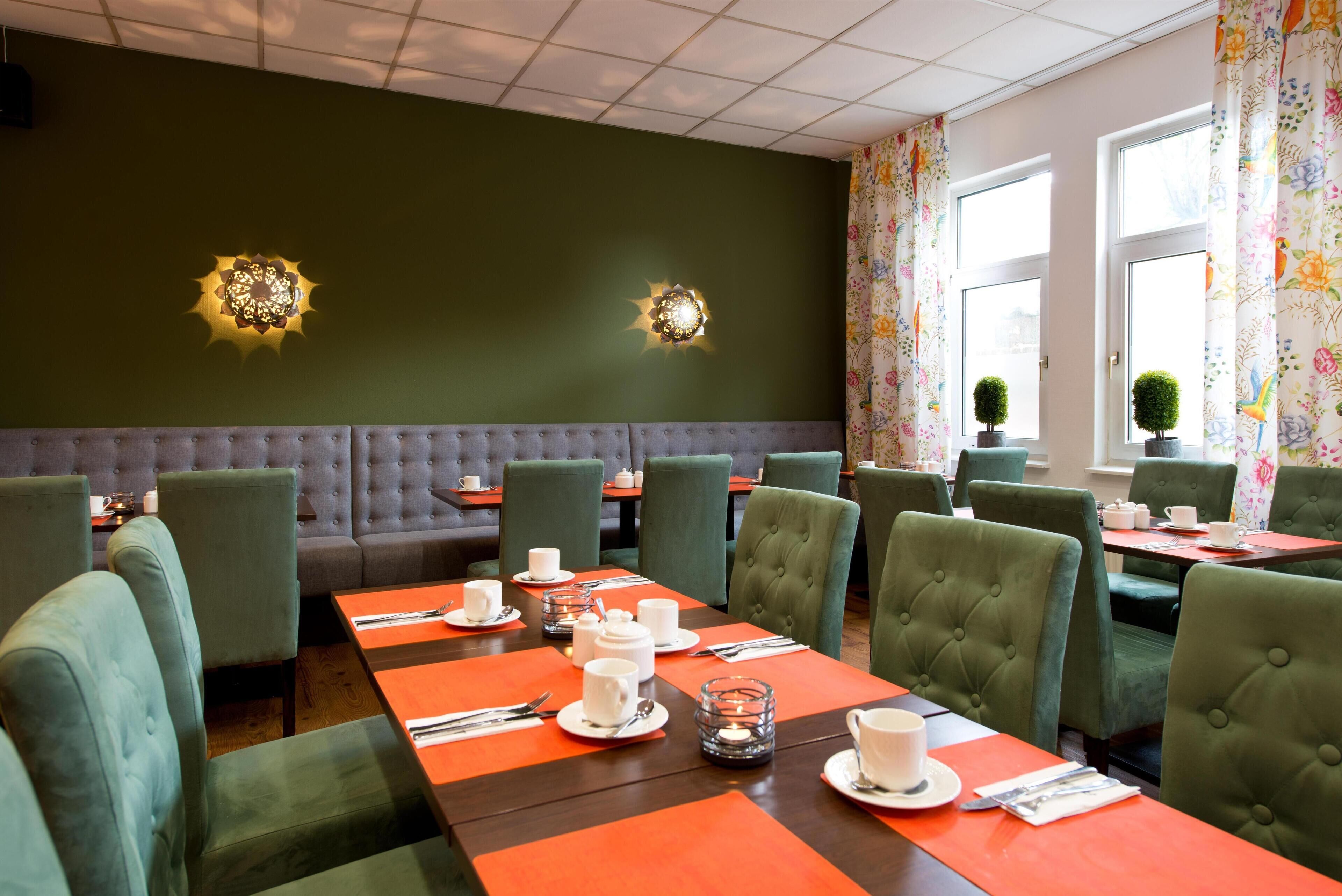 Un restaurante con asientos tipo banca tapizados en verde, mesas, apliques decorativos en la pared y cortinas florales.