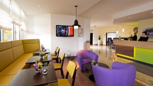 A modern hotel lobby with a reception desk, seating area with yellow and purple chairs, and a television mounted on the wall. - ACHAT Hotel Zwickau (Zwickau)