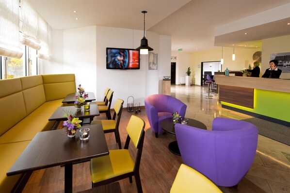 A modern hotel lobby with a reception desk, seating area with yellow and purple chairs, and a television mounted on the wall. - ACHAT Hotel Zwickau (Zwickau)