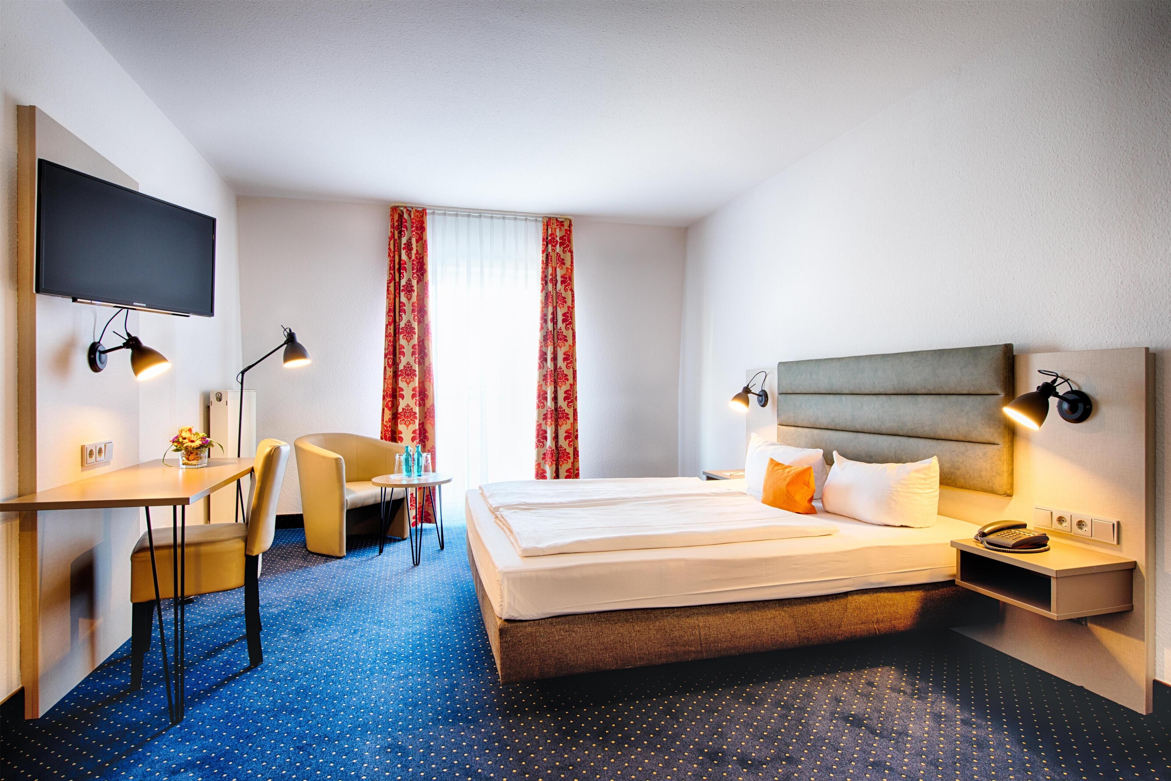 Una habitación de hotel con cama, escritorio, silla, televisor y ventana con cortinas.