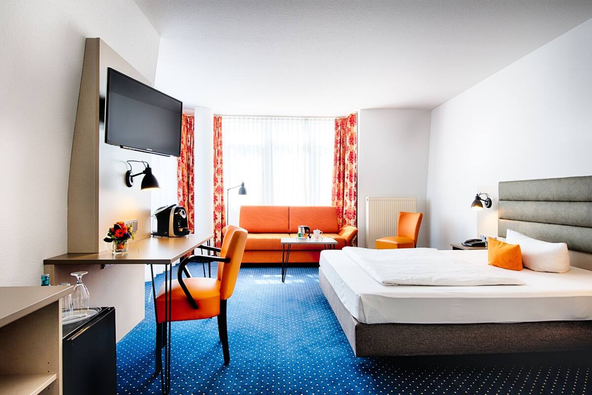 Une chambre d’hôtel avec un lit, un bureau avec une télévision, une chaise, un canapé et une fenêtre avec des rideaux.