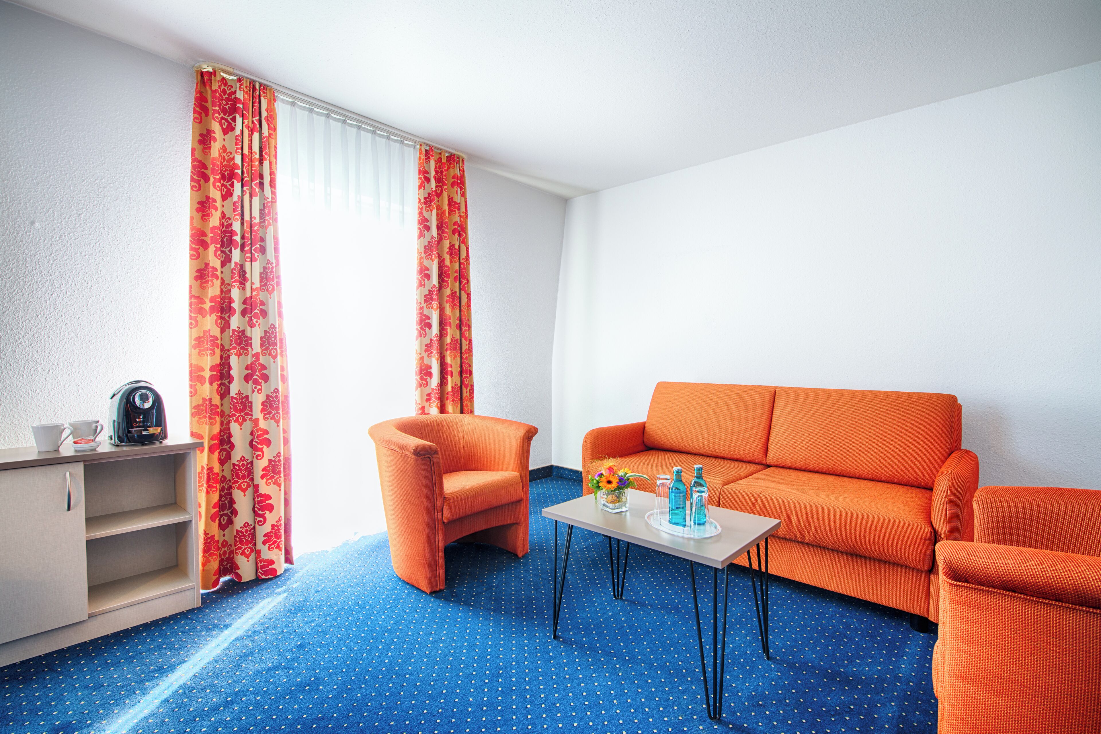 A room with a blue polka dot carpet, an orange sofa, a matching armchair, a small table with a tray, and a cabinet.