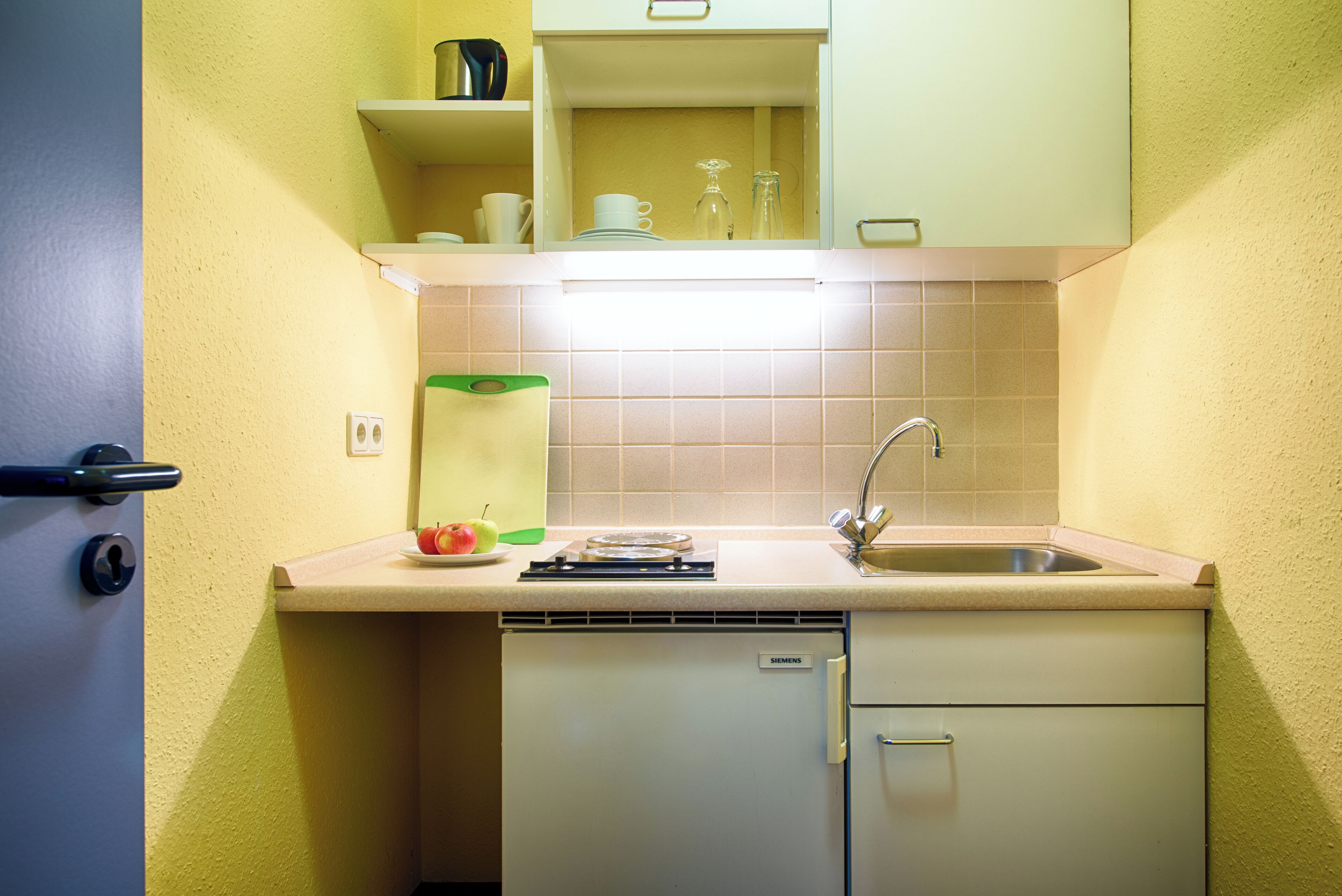 Cocina compacta con lavamanos, refrigerador Siemens y encimera con tabla de cortar y un plato de fruta.