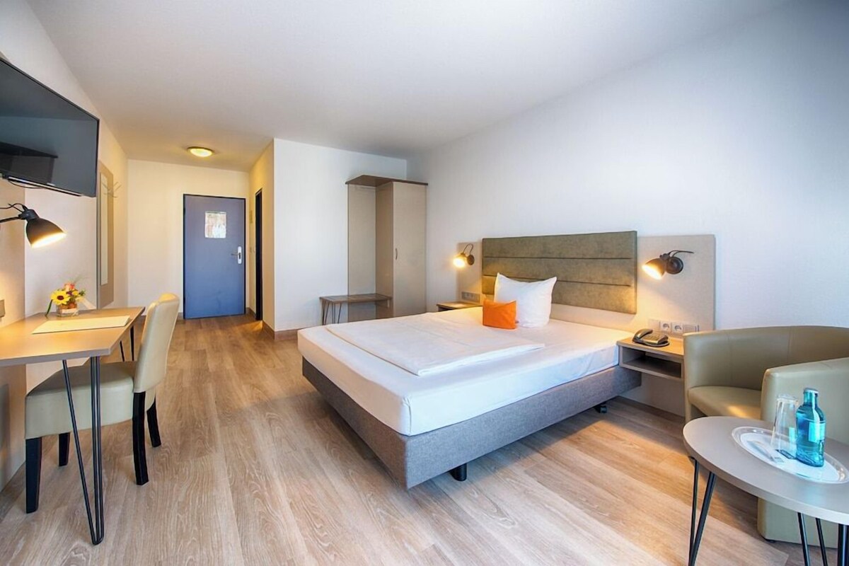 Una habitación de hotel moderna con cama, escritorio, silla, mesita de noche y una pequeña mesa con una botella.