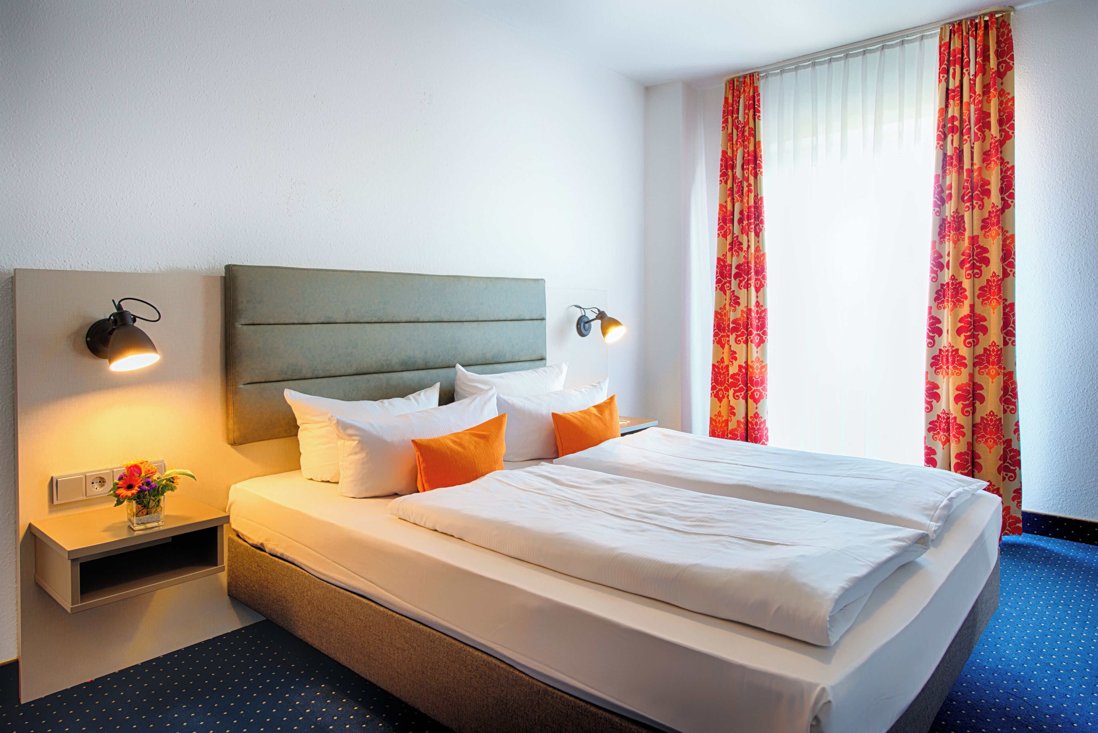 A double bed with white and orange bedding, a headboard, bedside lamps, and a nightstand with a flower vase.