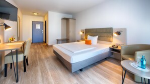 A modern hotel room with a bed, desk, chair, and a small table with a bottle. - ACHAT Hotel Zwickau (Zwickau)