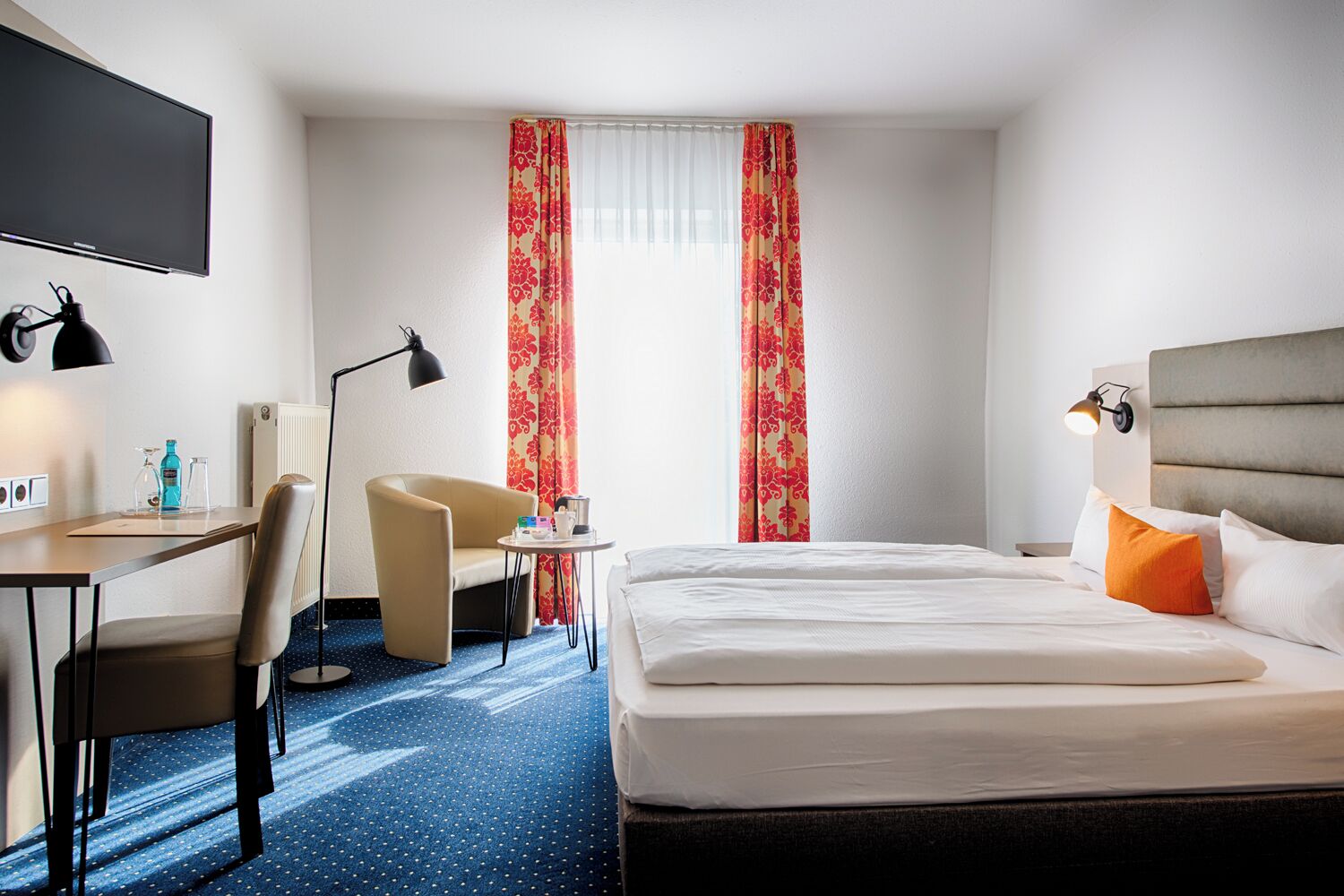 A hotel room with a bed, a desk with a chair, a small table, and a TV.