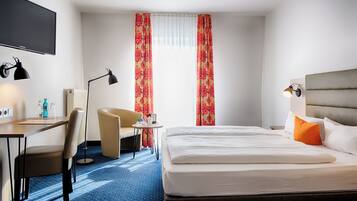 A hotel room with a bed, a desk with a chair, a small table, and a TV.