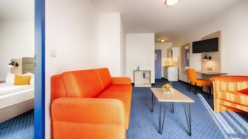 A hotel room with a blue carpet, orange sofa, a small table, and a TV mounted on the wall.