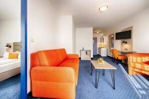 A hotel room with a blue carpet, orange sofa, a small table, and a TV mounted on the wall.