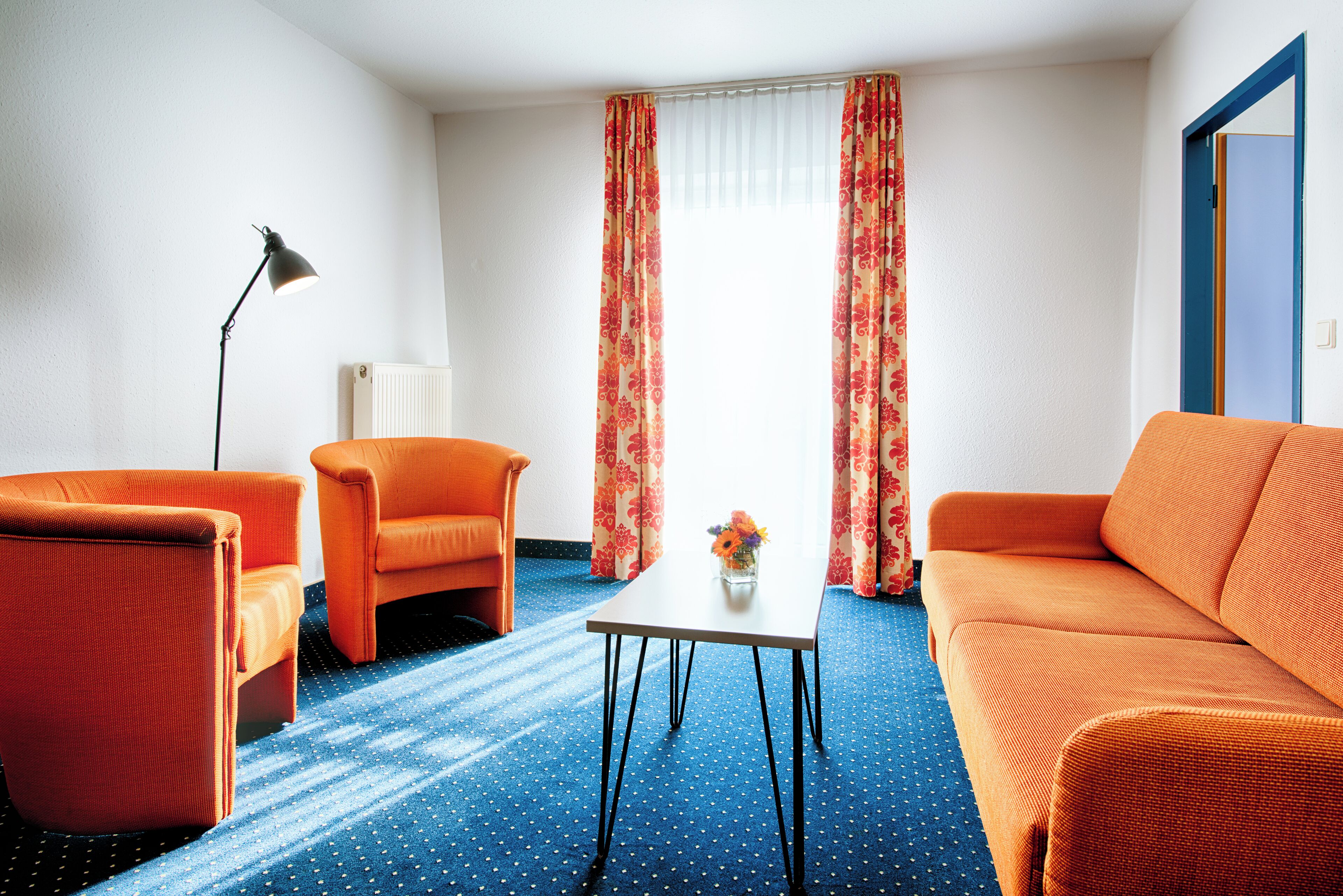 Een kamer met een blauw tapijt, oranje meubels en een raam met rode gordijnen.